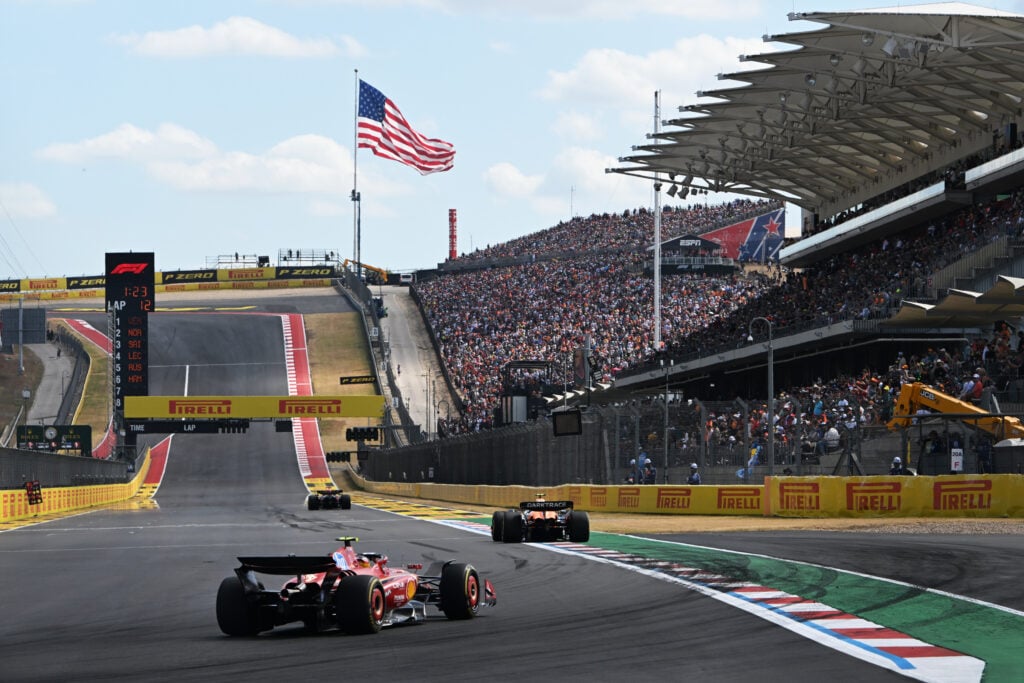 Formula 1 cars head round the final corner along the main straight and past the grandstands at the Circuit of the Americas. Turn 1 and a big American Flag sit off in the distance