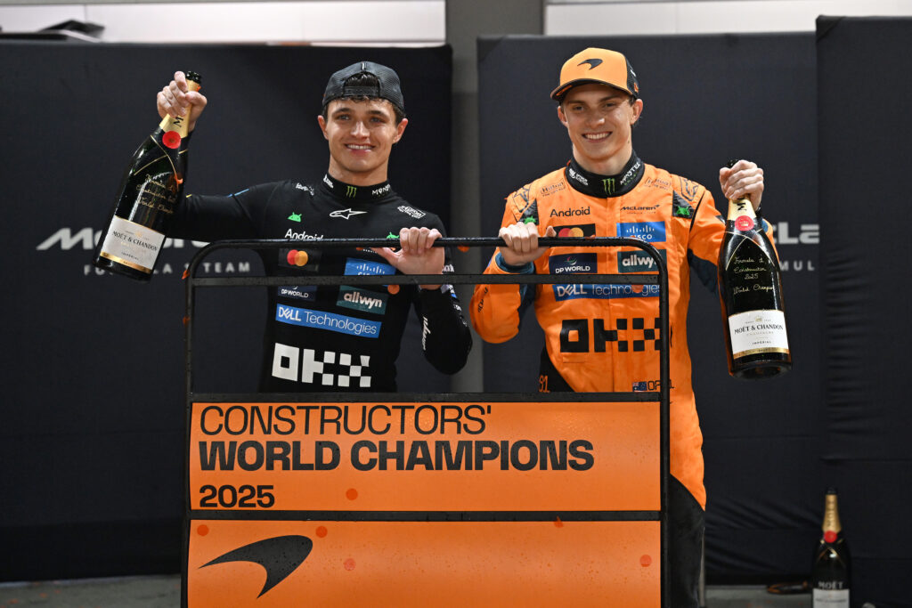 Lando Norris and Oscar Piastri of McLaren smile as they hold up bottles of champagne and a pit wall board which reads "constructors' world champions 2025" and includes a team logo